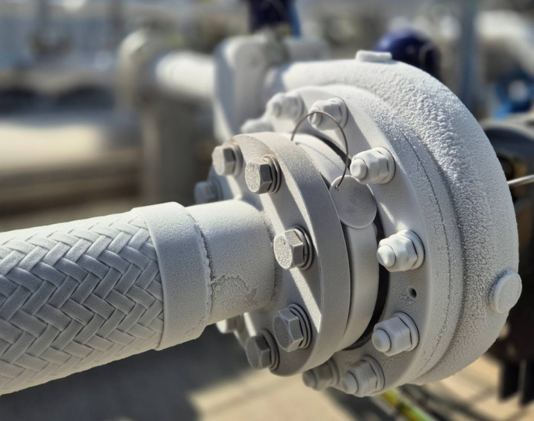 Närbild av en industriell rör- och flänsanslutning med metallflätad slang, helt täckt av ett vitt lager rimfrost på grund av extrem kyla av kryogen gas.