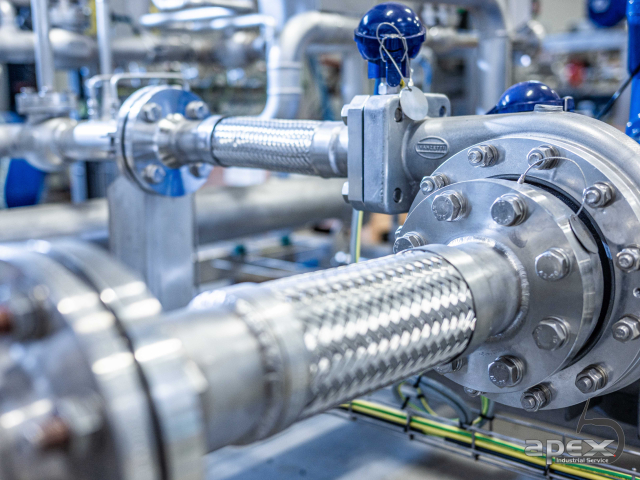 Industriell rörinstallation med stålomspunna flexslangar, rörflänsar och Vanzetti-pumpenhet från Apex Industrial Service.